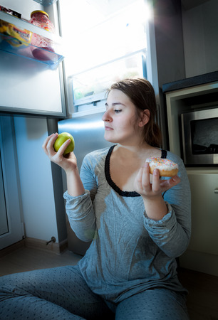 Portrait of woman holding apple and donut against fridge at nightの写真素材