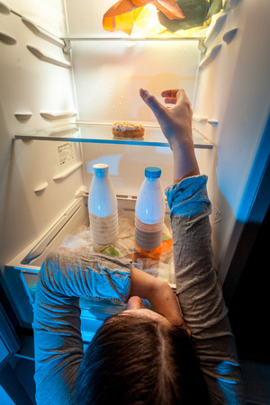 Young woman taking donut from top shelf of refrigeratorの写真素材