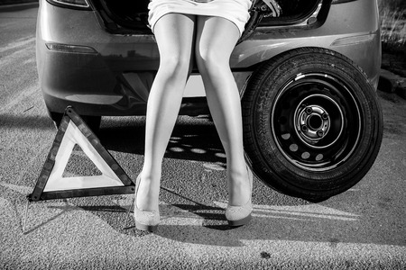 Black and white closeup photo of woman sitting on broken car near warning sign and spare wheelの写真素材