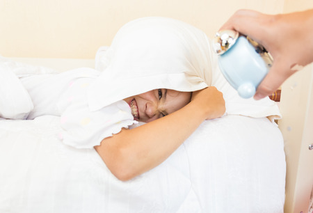 Little girl covering head with pillow and being awakened by alarm clockの写真素材