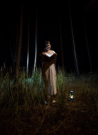 Young woman in nightgown reading book at night forestの写真素材