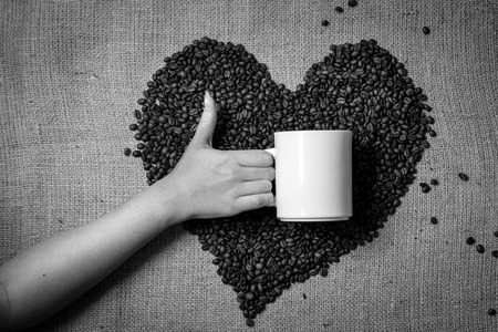 Black and white photo of hand with thumb up holding mug against heart made of coffee beansの写真素材