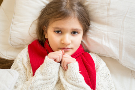 Portrait of little sad girl in white sweater lying under blanket at bedの写真素材