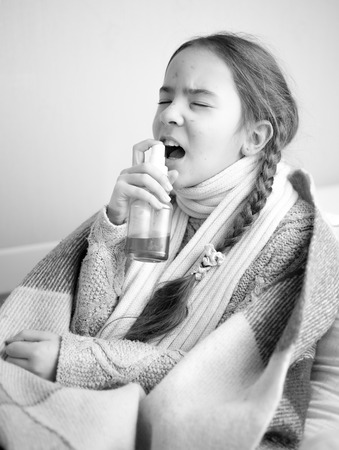 Black and white portrait of little girl lying in bed and using throat sprayの写真素材