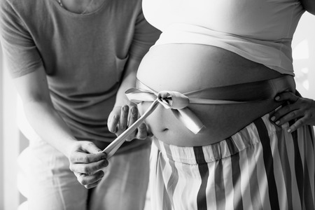 Black and white closeup photo of young man untying ribbon on wife's pregnant abdomen