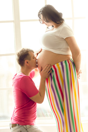 Portrait of kneeling man kissing pregnant wife's belly at living roomの写真素材