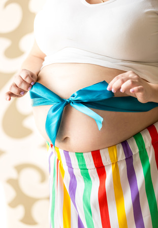 Closeup photo of cute pregnant woman tying bow on stomachの写真素材