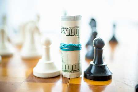 Closeup conceptual photo of twisted dollar bills standing on chess board between white and black piecesの写真素材