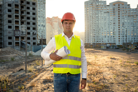 Portrait of young engineer posing with blueprints against building site