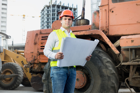 Portrait of architect reading blueprints next to bulldozer at building site