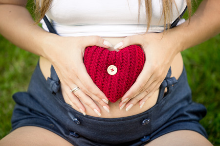 Closeup photo of pregnant woman holding red knitted heart at stomachの写真素材