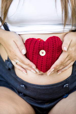Conceptual photo of caring expecting mother holding red heart at abdomenの写真素材
