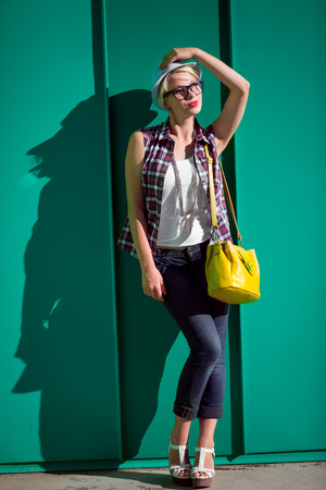 Full length portrait of stylish hipster girl leaning against blue wall on street at sunny dayの写真素材
