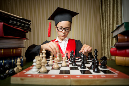 Portrait of genius girl in graduation cap playing chess with herself
