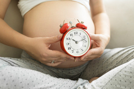 Closeup photo of pregnant woman holding red alarm clock at tummyの写真素材