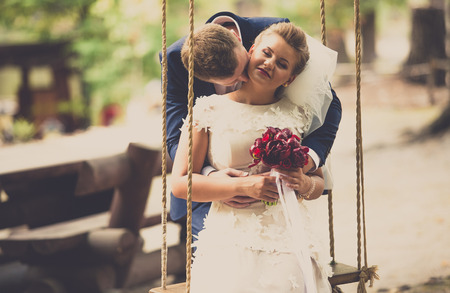 Toned portrait of young groom kissing bride sitting on swing at parkの写真素材