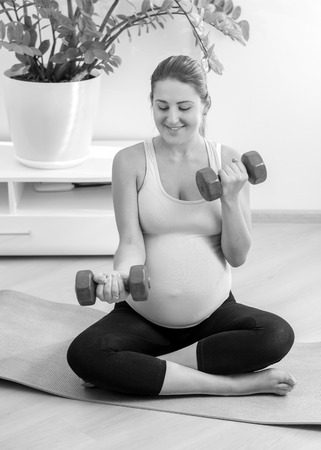Black and white photo of pregnant woman exercising with dumbbells at homeの写真素材