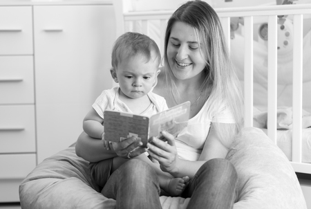 Black and white portrait of young cheerful mother sitting with baby in beanbag and reading bookの写真素材