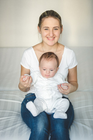 Portrait of beautiful smiling mother sitting on bed and holding her baby boyの写真素材