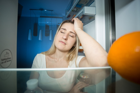 Portrait of sleepy woman leaning on refrigerator shelf and trying to sleep