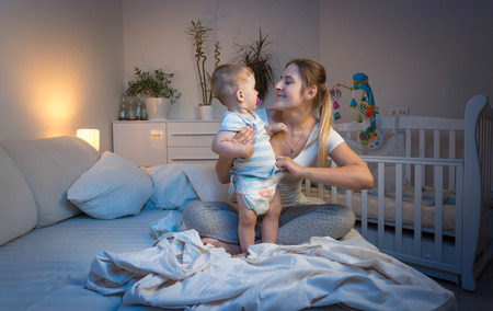 Cute cheerful baby boy playing with mother on bed at night