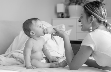 Black and white photo of beautiful smiling mother feeding her 9 months old baby from bottle at bedroomの写真素材
