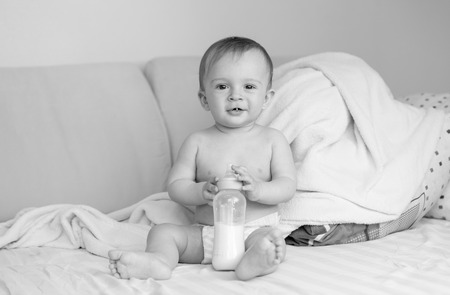 Black and white photo of cute baby boy sitting on bed with milk bottleの写真素材