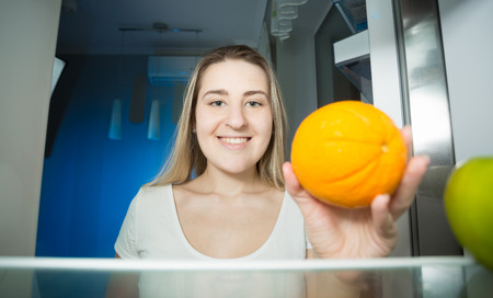 Portrait of beautiful smiling woman holding orange and looking inside the refrigeratorの写真素材