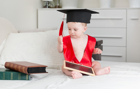 Cute 10 months old baby boy in graduation cap holding digital tablet and looking at stack of booksの写真素材