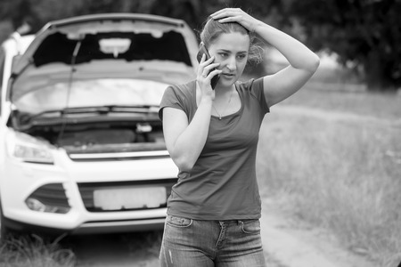Black and white portrait of sad woman standing at broken car and calling for helpの写真素材