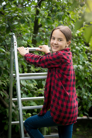 Portrait of beautiful smiling teenage girl in red checkered shirt climbing up stepladder at garden
