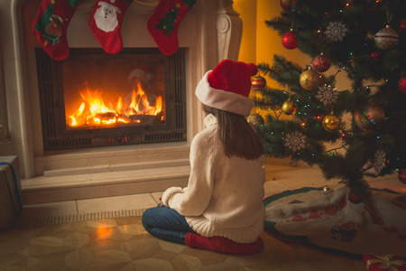 Toned image of beautiful girl in Santa hat sitting on floor and looking at fireplaceの写真素材