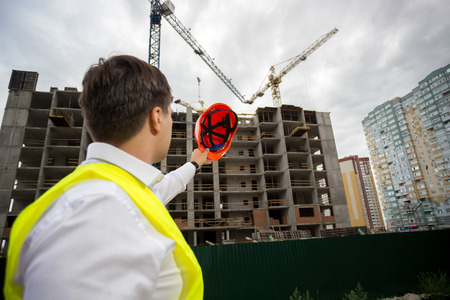 Young construction engineer pointing at building under construction with red hardhat