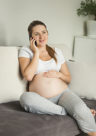 Beautiful pregnant woman sitting on sofa and talking by phoneの写真素材