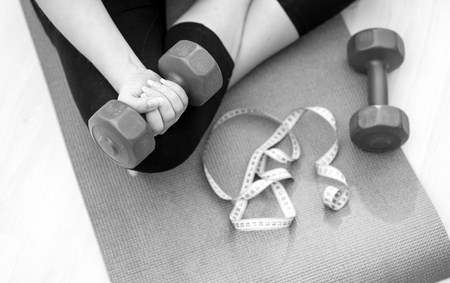 Closeup black and white photo of young woman sitting on fitness mat and lifting dumbbellsの写真素材