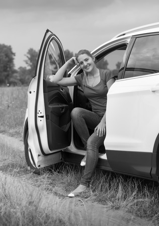 Black and white image of beautiful smiling woman sitting on car drivers seat at fieldの写真素材