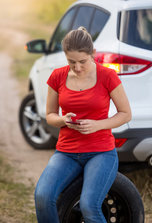 Woman sitting on spare tire next to broken car at field and searching for service phone numberの写真素材