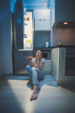 Sleepy woman sitting on floor at open fridge at late nightの写真素材