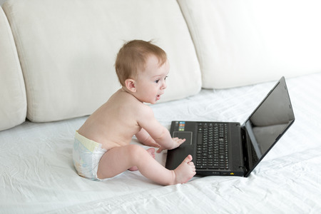 Baby boy in diapers sitting on bed and typing on laptopの写真素材