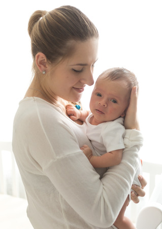 Portrait of beautiful young mother holding newborn baby at big windowの写真素材