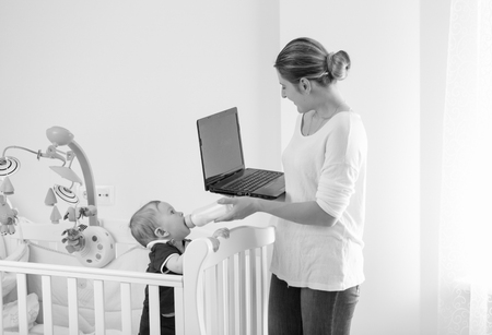 Black and white image of young businesswoman working at home and giving milk to her babyの写真素材