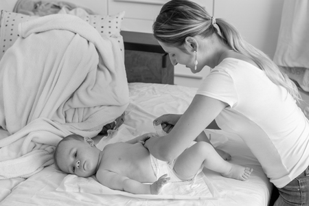Black and white portrait of happy young mother changing her baby's diapers on bedの写真素材