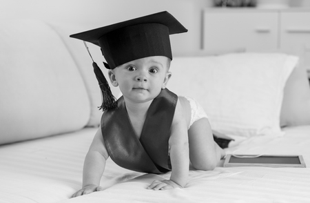 Black and white image of adorable 10 months old baby boy in graduation cap crawling on bedの写真素材