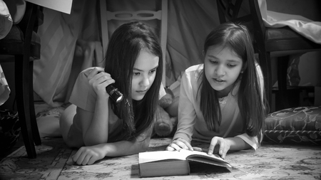 Black and white portrait of two teenage girls lying on floor and reading book with flashlightの写真素材
