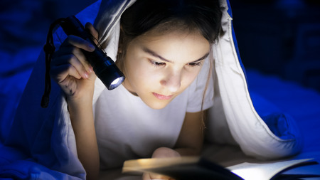 Portrait of teenage girl reading book with flashlight at nightの写真素材