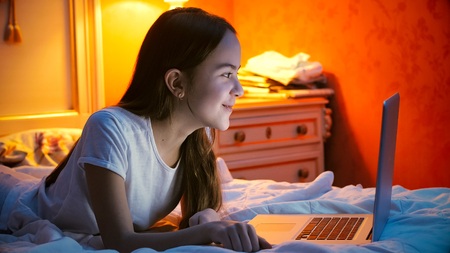 Portrait of beautiful smiling girl lying on bed with laptop before going to sleepの写真素材