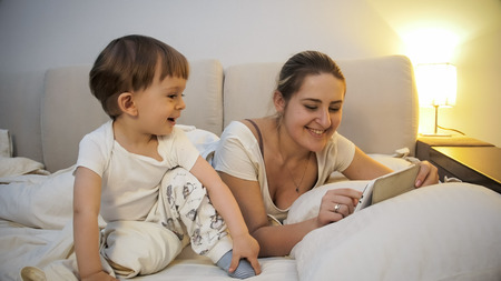 Portrait of smiling young mother with toddler son using digital tablet computer in bed
