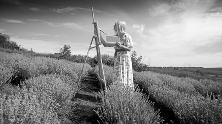 Black and white image of young woman in long dress painting picture of fieldの写真素材