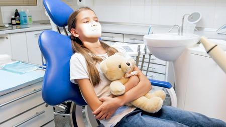 Cute girl with teddy bear sitting in dentist chair in clinic and feeling nervous before treating teethの写真素材