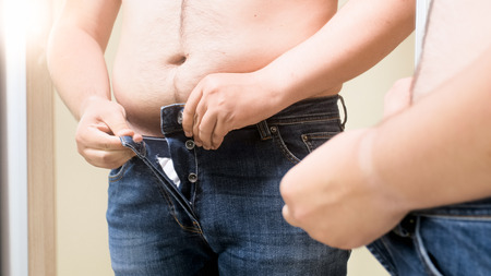 Closeup photo of young fat man buttoning tight jeans at mirrorの写真素材